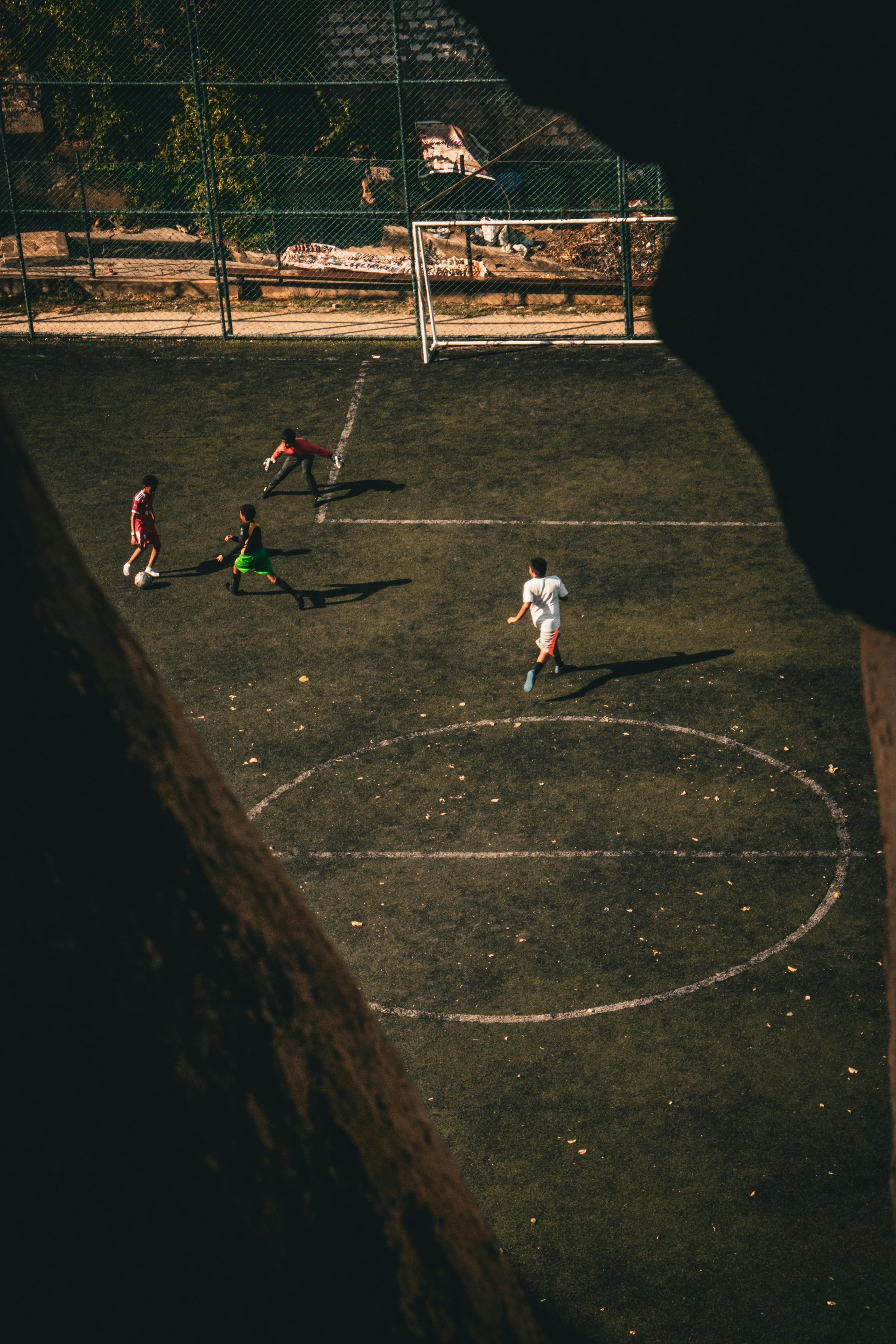 Four players on a small outdoor soccer court, with a goal at the top in dim light.
