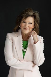 A woman in a pink jacket and green shirt is standing with her arms crossed.