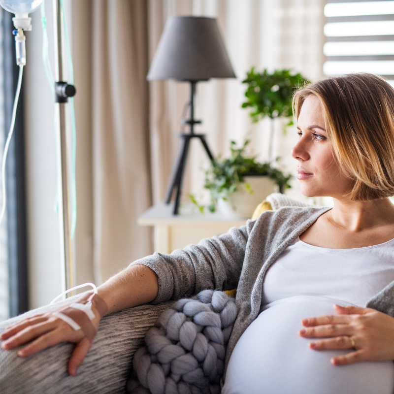 Pregnant person receiving IV, seated on couch, looking out window, light and airy interior.