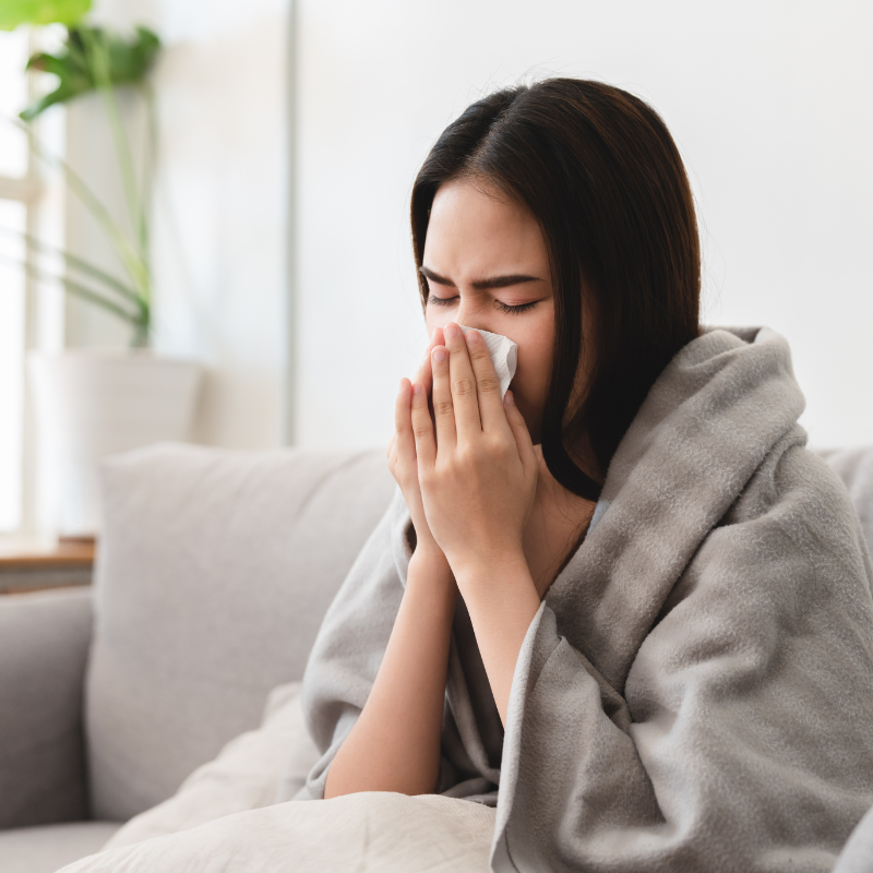 Woman wrapped in blanket blowing nose with tissue on a couch.