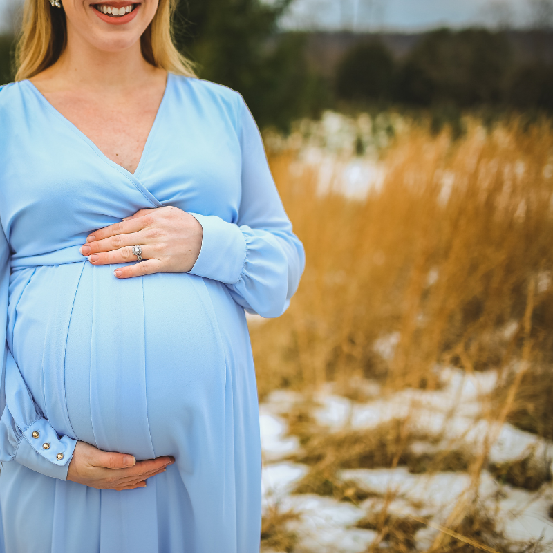 Pregnant person in a blue dress, cradling belly in a field with tall grass and snow.