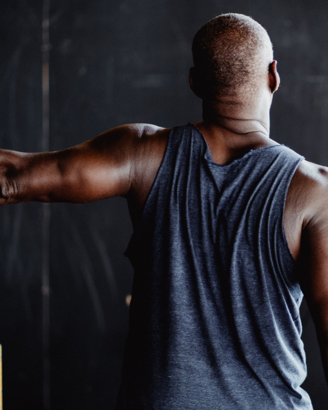 Man in blue tank top, arms outstretched, exercising in front of a dark background.