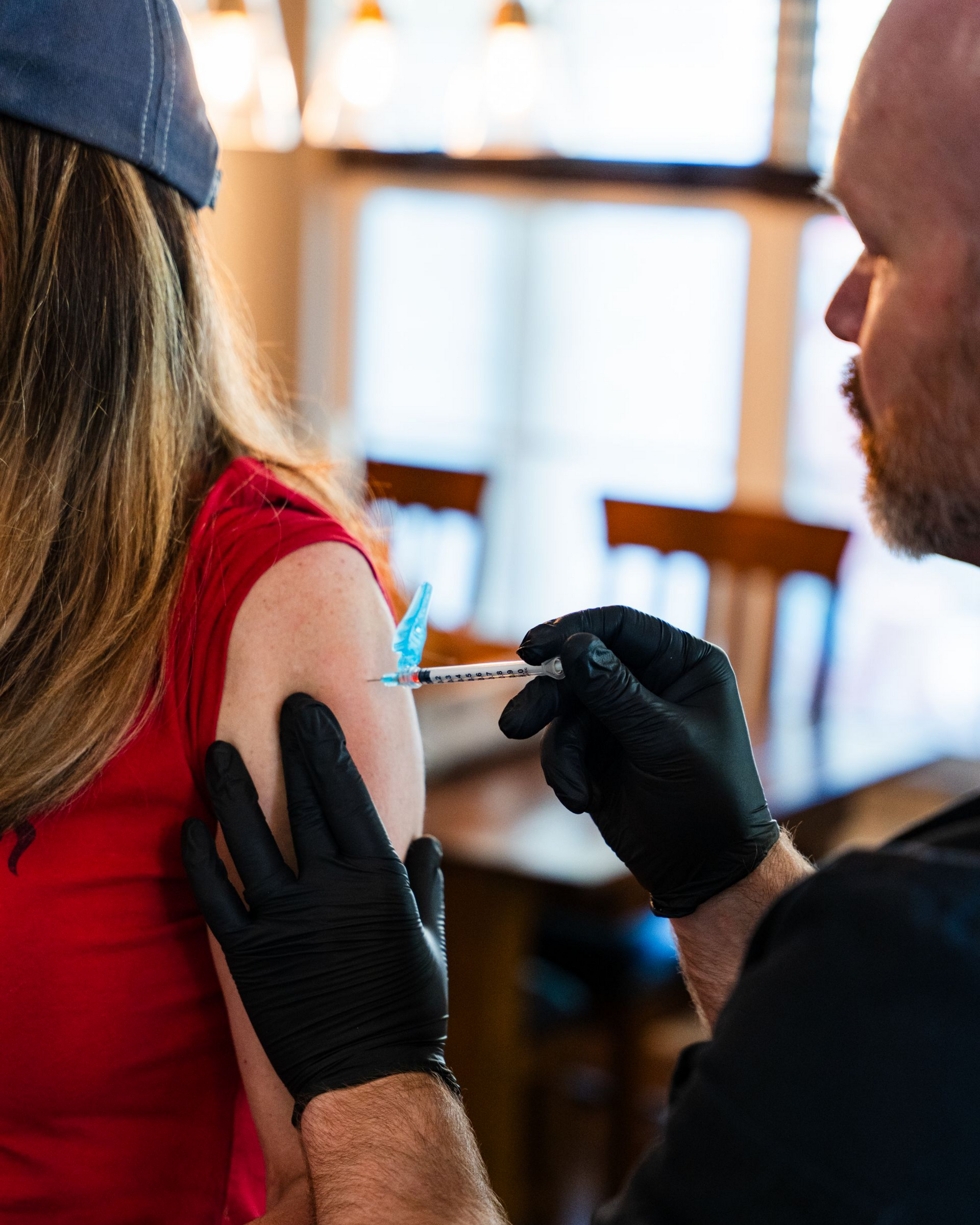 Person in red shirt receiving injection in arm from person in black gloves. Interior setting, blurred background.