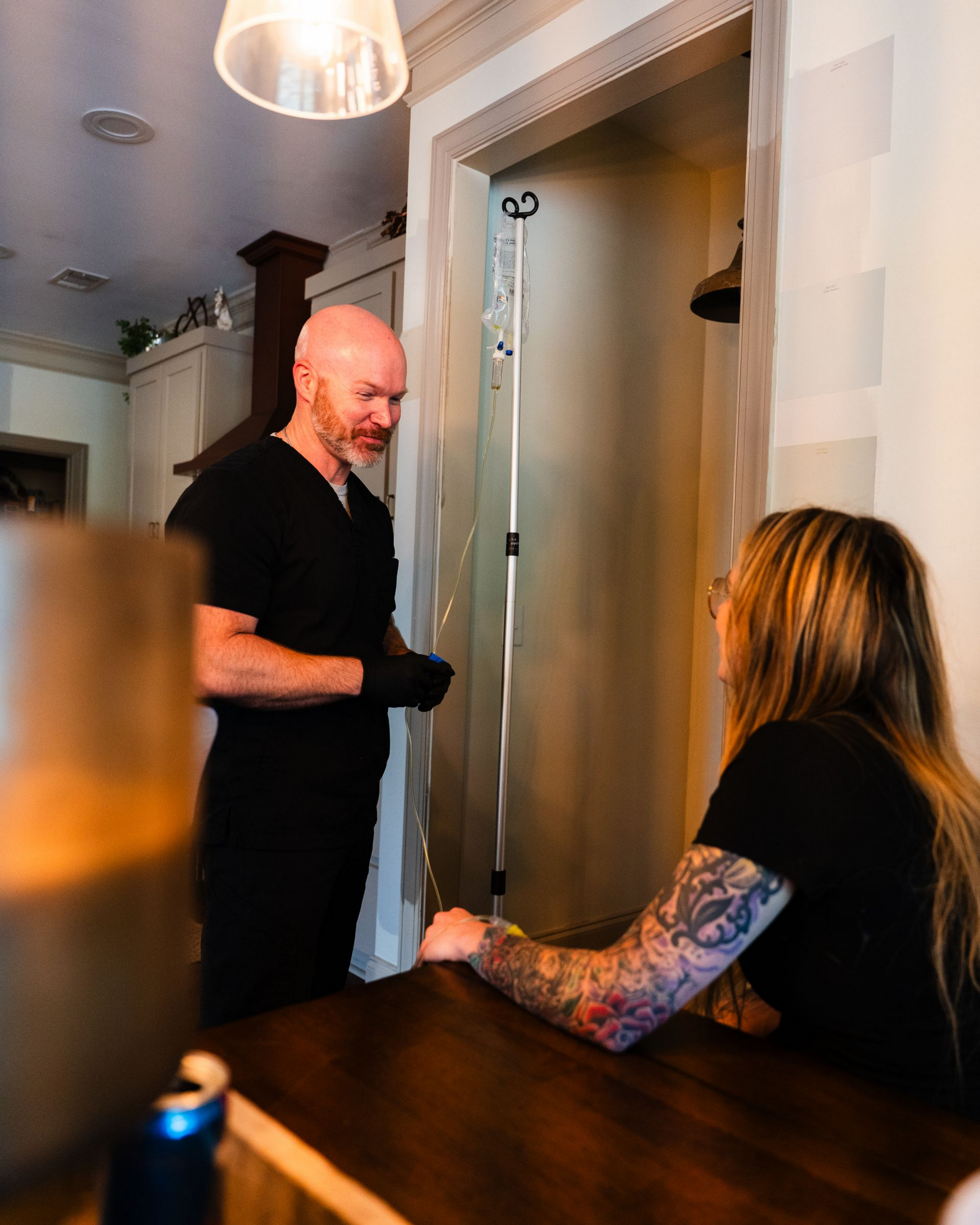 Man in black scrubs prepares IV drip for woman with arm tattoo in a doorway.