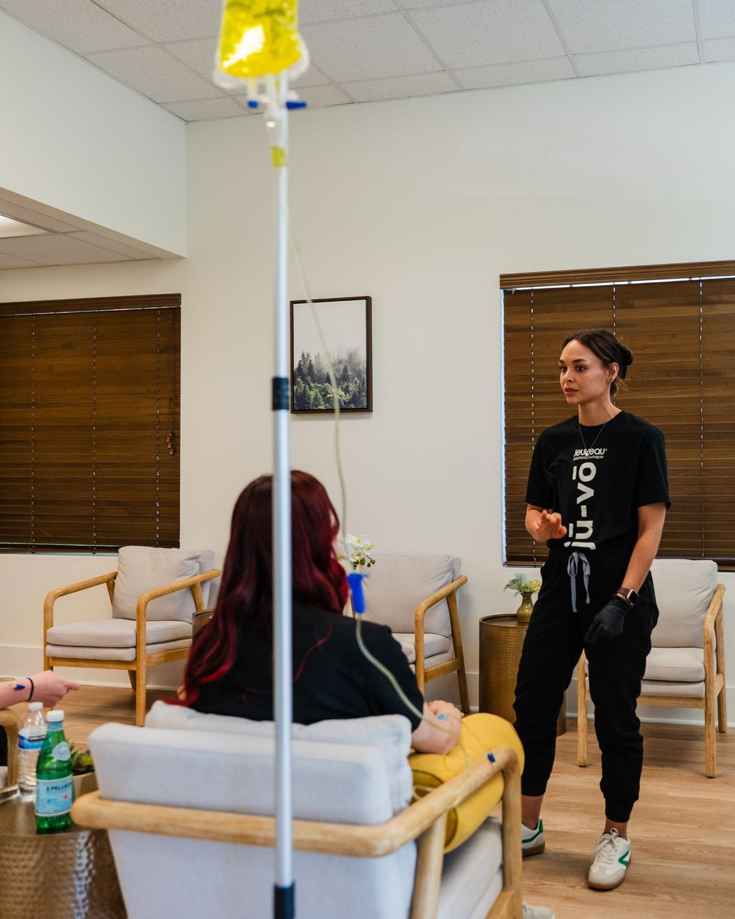 Woman receiving IV drip in a clinic; staff member explains procedure. Bright room with comfortable seating.