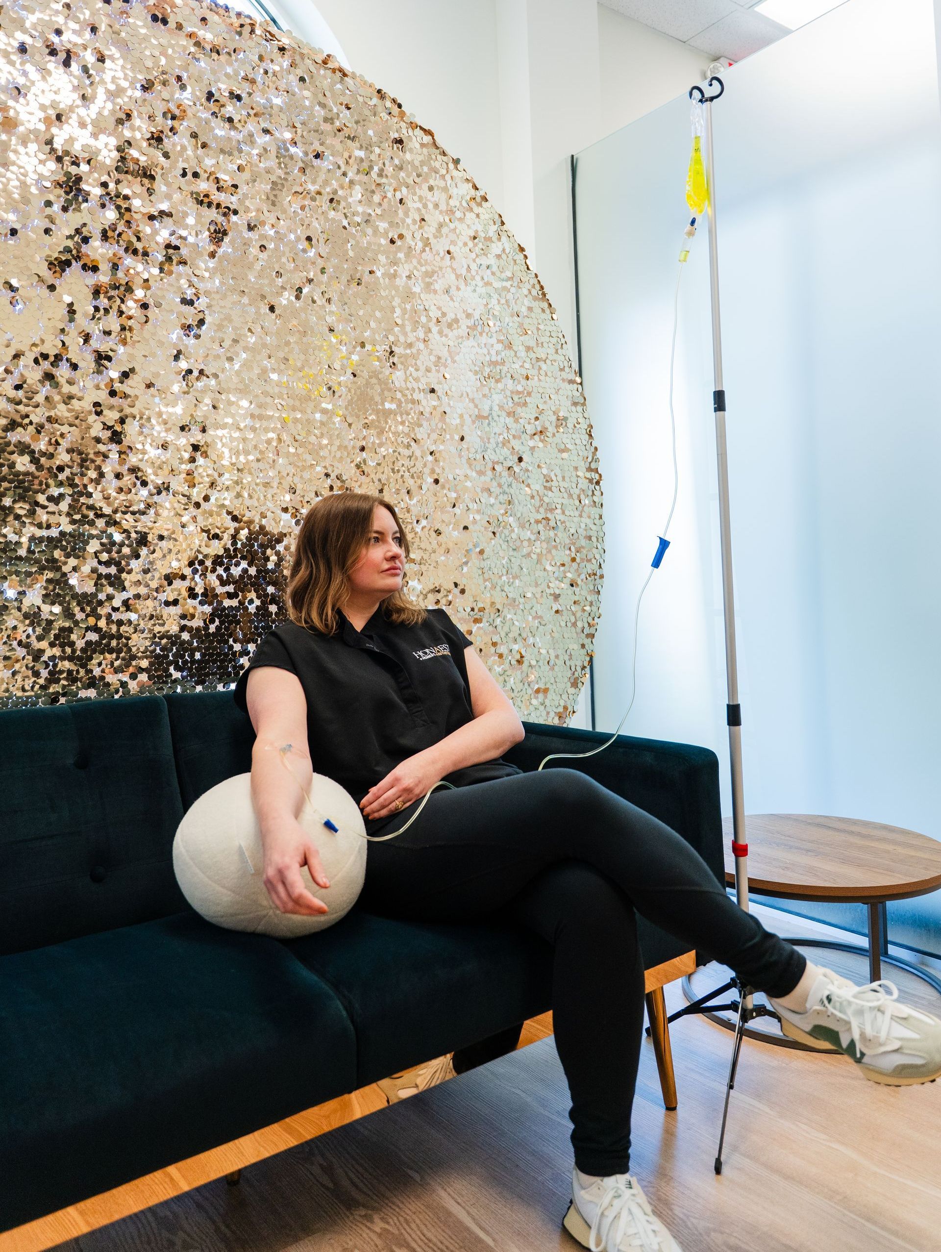 Woman receiving IV drip while sitting on a couch, with a gold sequin backdrop.