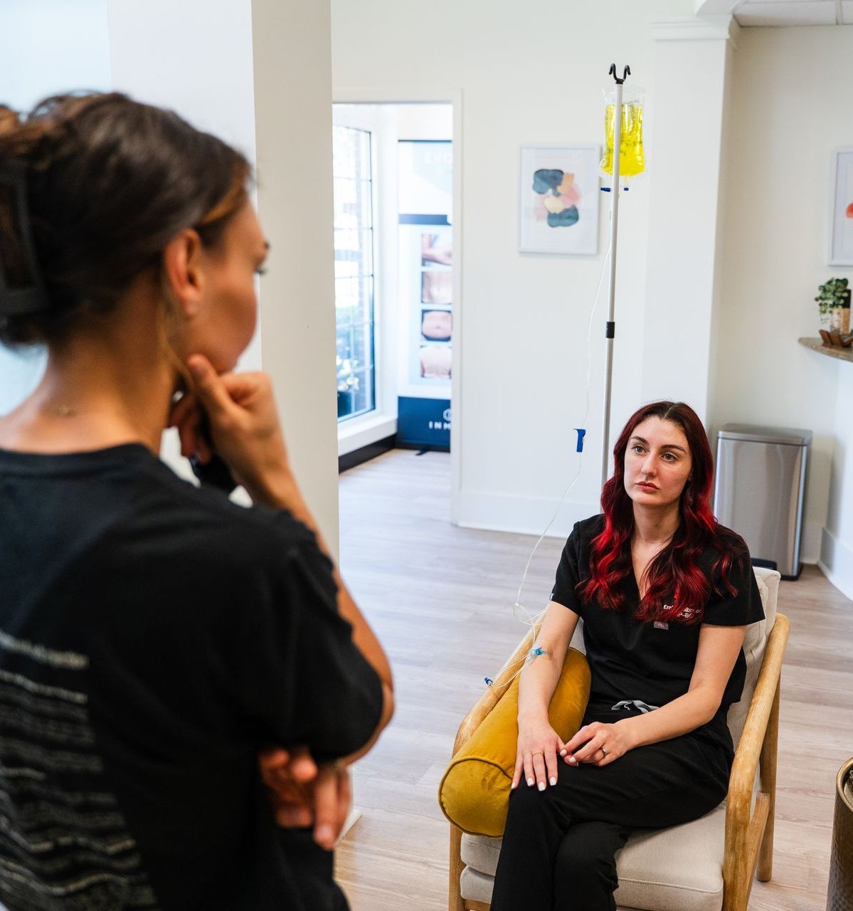 Woman in black shirt speaks with nurse with red hair, IV bag in background.