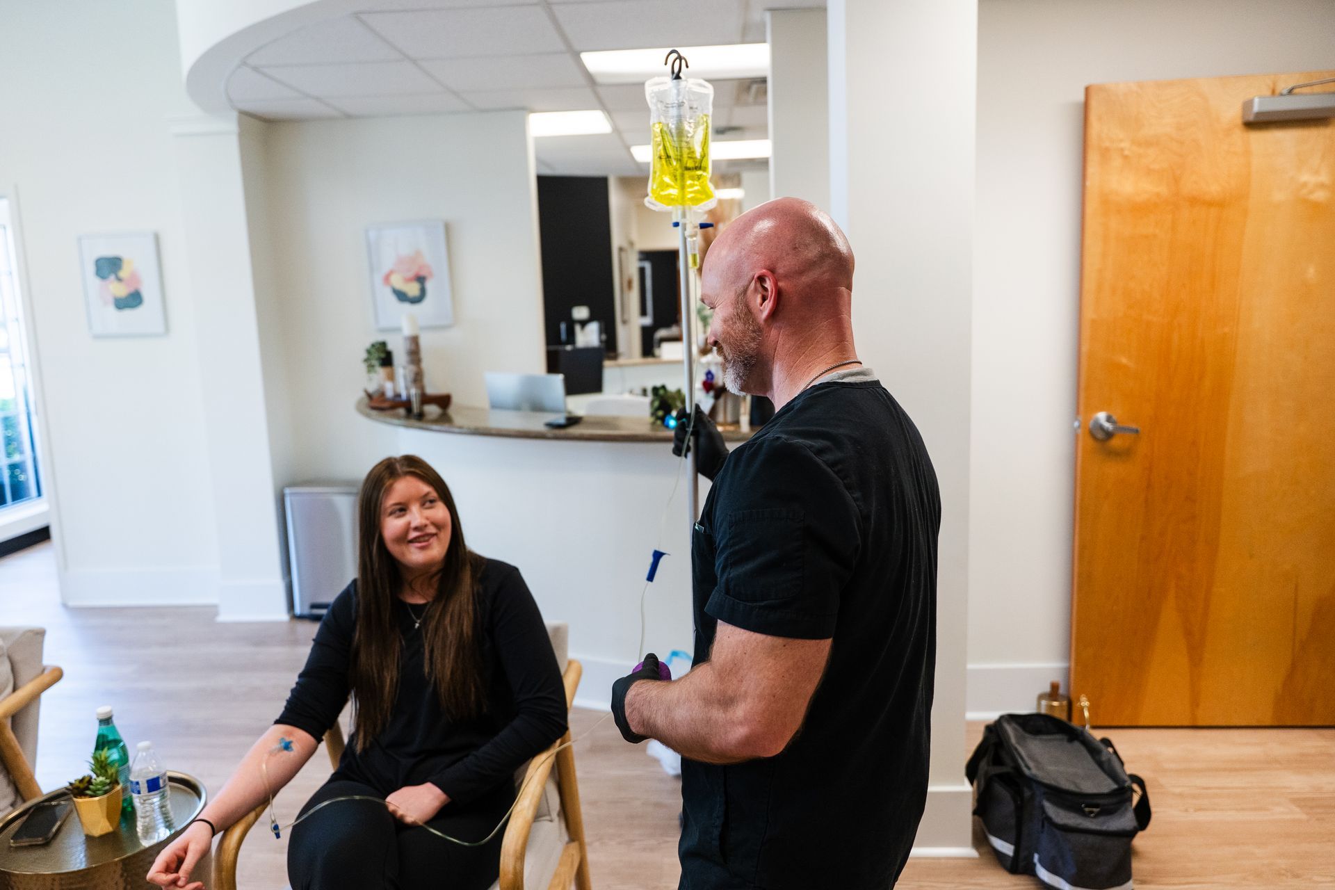 Man in black scrubs laughs, facing a person in a room with a window, chair, and blinds.