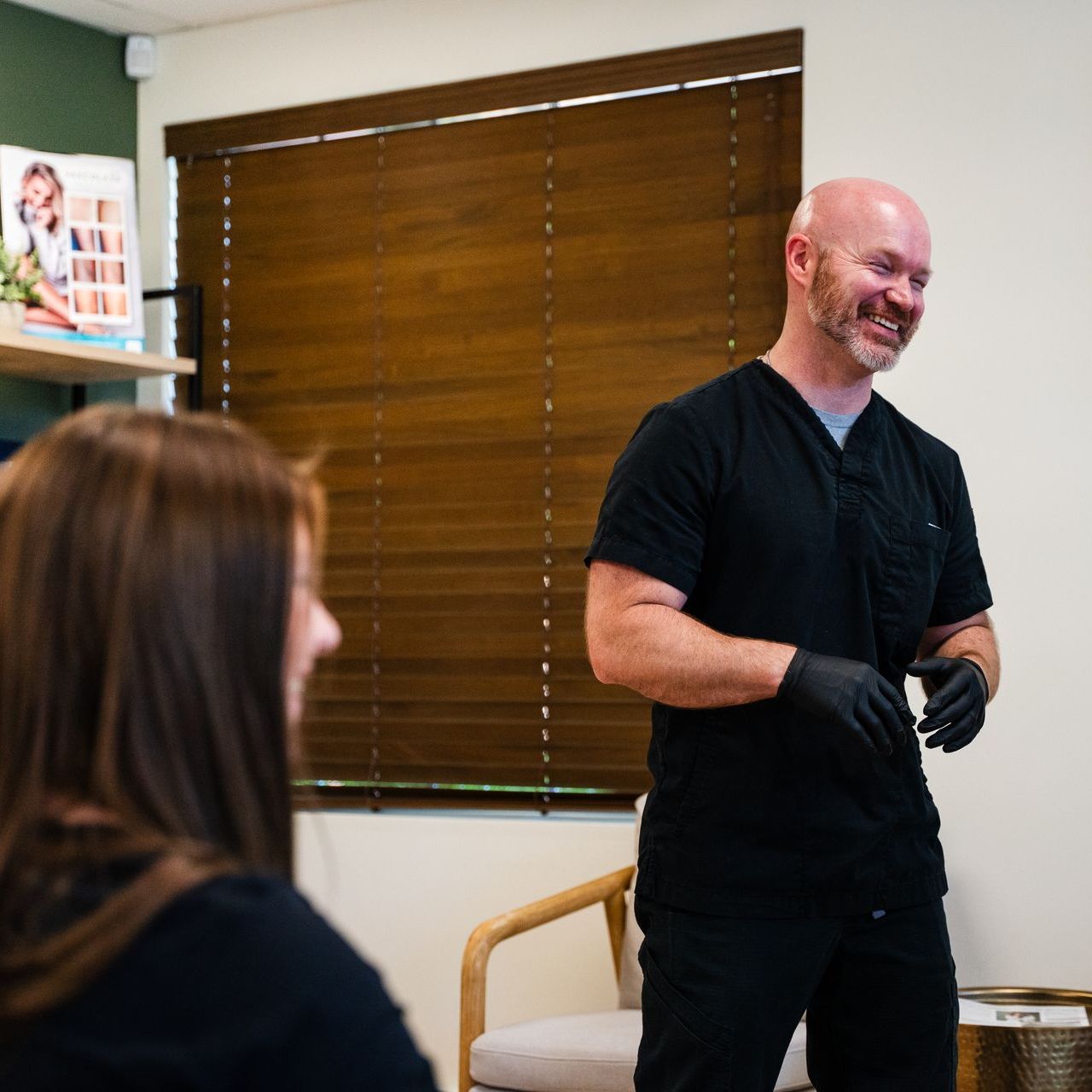 Man in black scrubs laughs, facing a seated person; office setting with blinds.