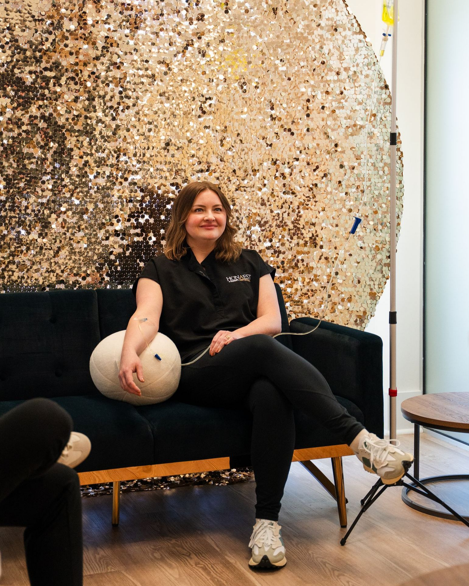 Woman in black shirt and leggings sits on a blue couch, smiling, in front of a gold sequined backdrop.