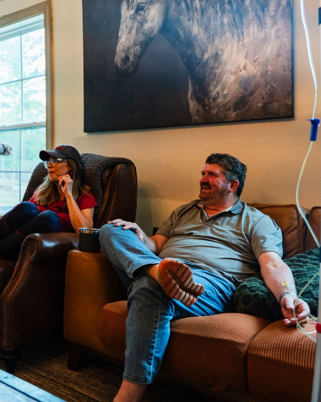 Couple relaxing on leather couches indoors, one with an IV drip. Horse art on the wall.