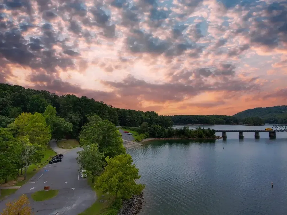 Lake scene at sunset: bridge, trees, vehicles on shore, and colorful cloudy sky.