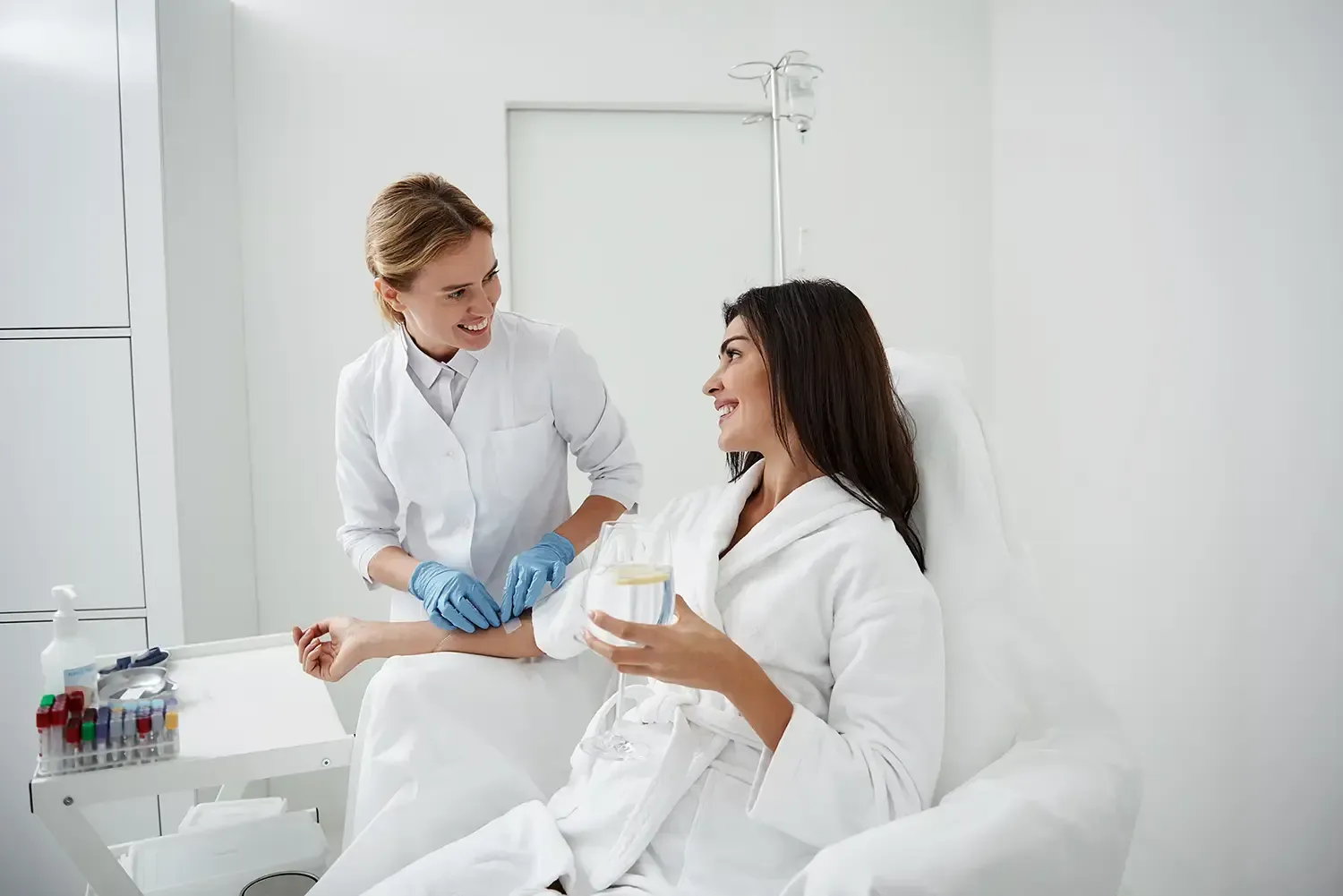 Woman receiving IV treatment; nurse prepares arm. Bright room, white coat, gloves, smiling.