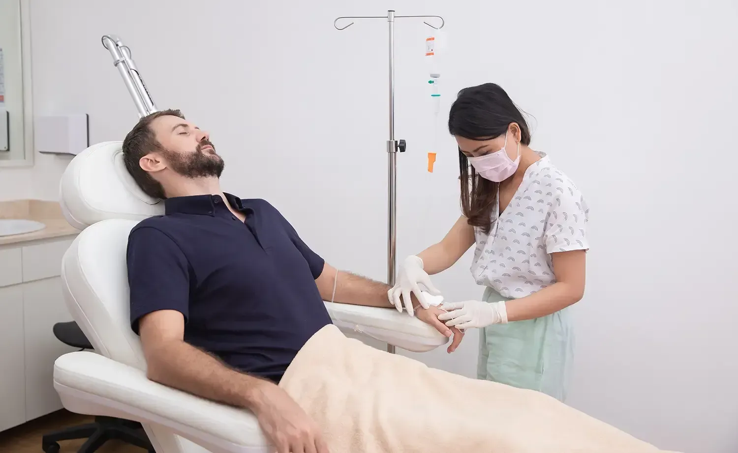 Man receiving IV treatment, attended by a masked nurse in a medical setting.