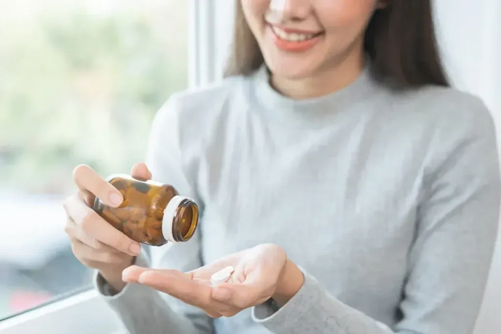 Woman smiling, pouring pills from a brown bottle into her hand near a window.