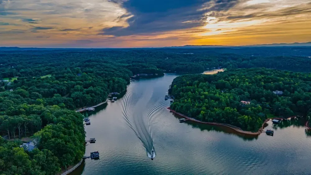 A boat travels through a winding lake surrounded by lush green trees under a colorful sunset.