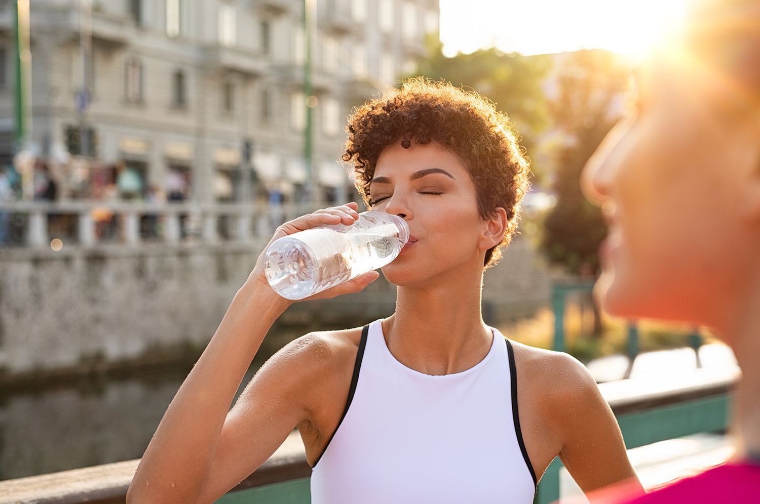A person in a white workout top drinks from a clear plastic water bottle outdoors in the sunlight.