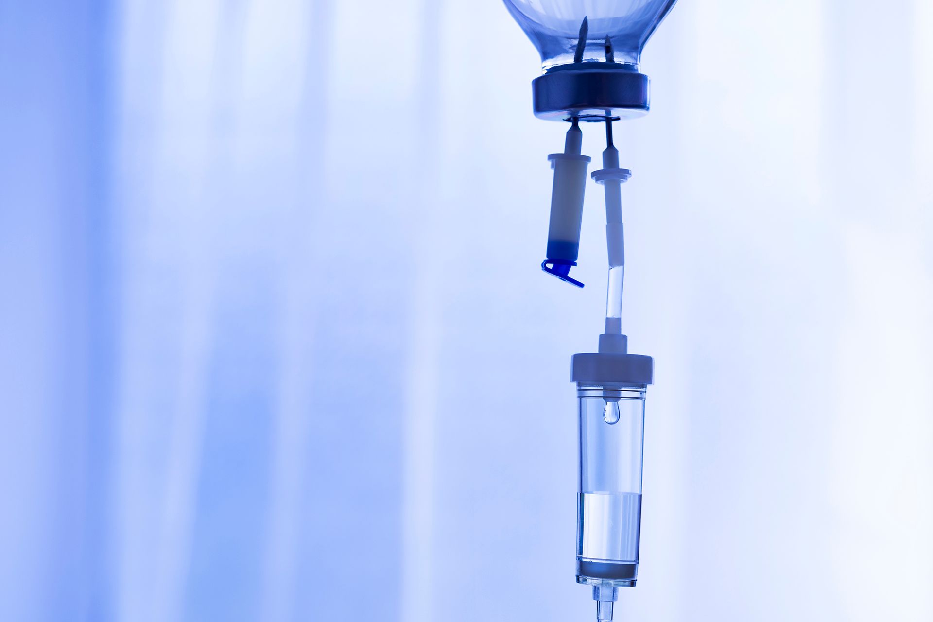 Close-up of a blue-tinted medical IV drip setup with a fluid bag, drip chamber, and tubing against a blurred background.