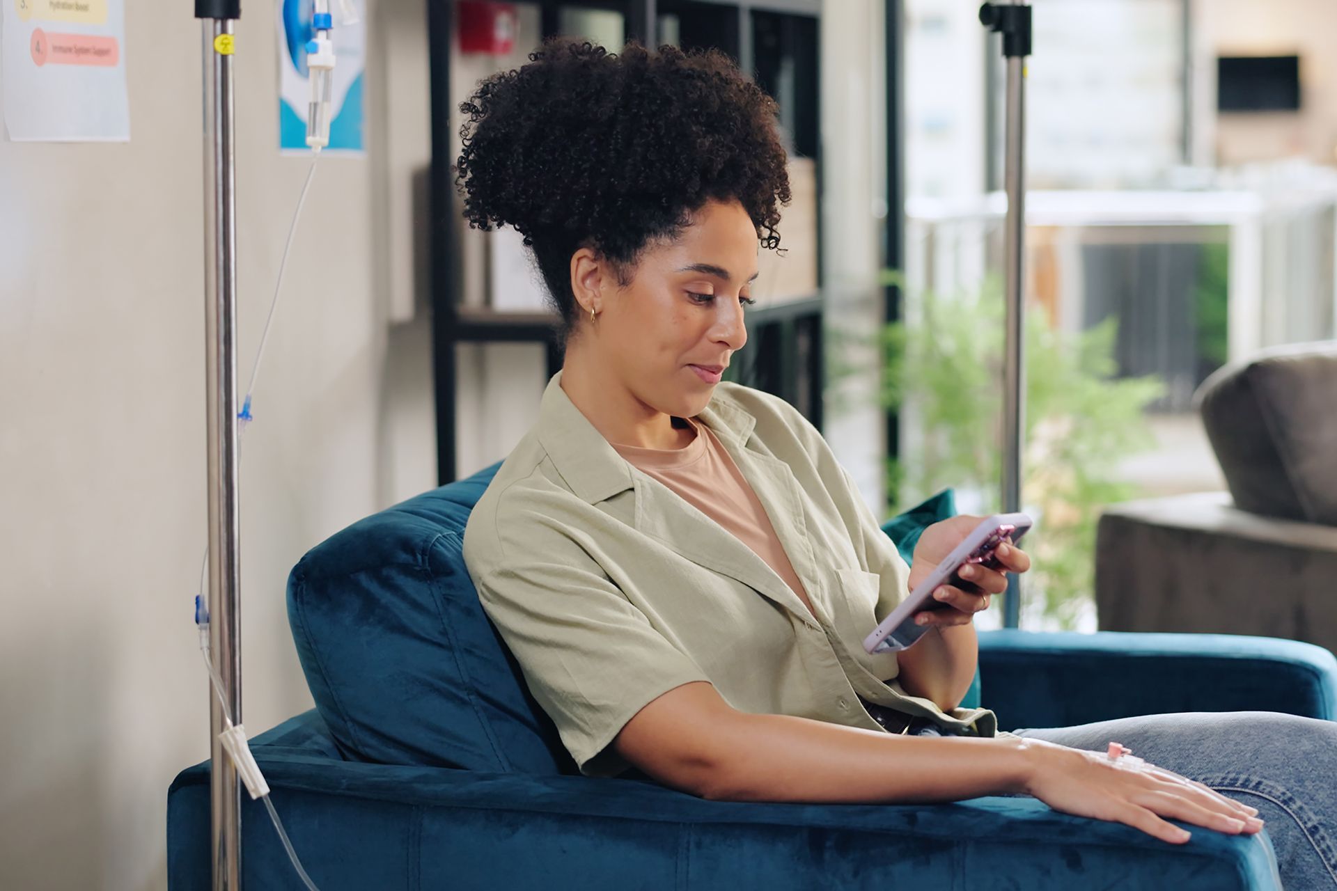 A person sits in a blue armchair in a medical setting, smiling while looking at a smartphone held in their hands.