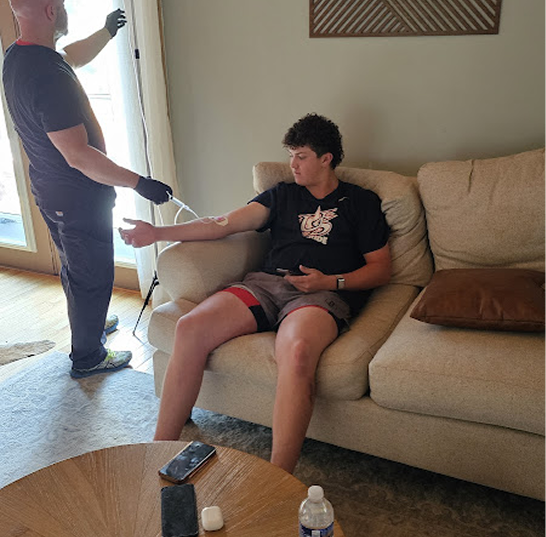 Man receiving an IV infusion while seated on a couch. Another person is administering the IV, indoors.