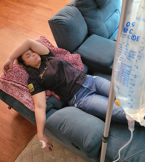 Person resting on a blue couch, receiving IV fluids. Dark shirt, jeans, and a pink blanket.