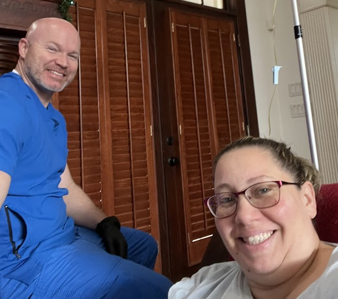 A person in blue scrubs and another person smile indoors, near a wooden door.