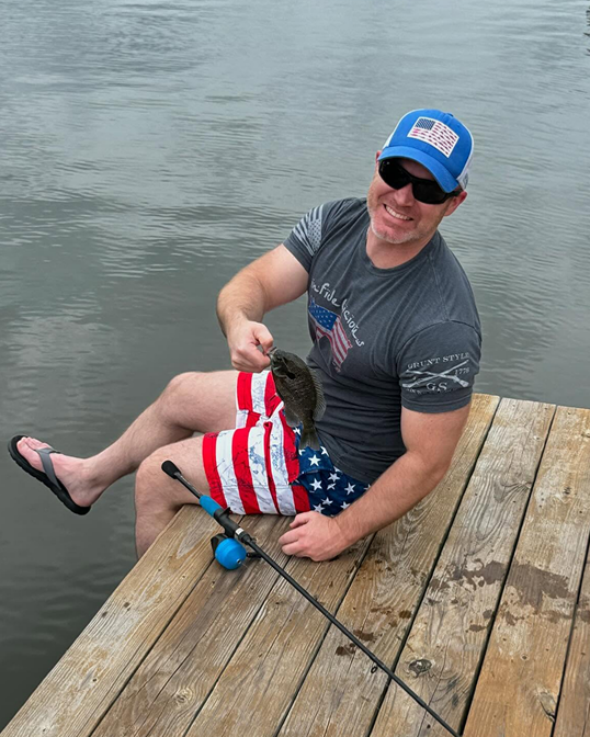 Man on a dock holding a small fish, wearing American flag attire. Water in background.