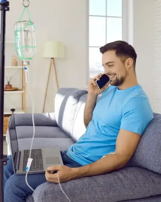 Man on sofa with IV drip, talking on phone, using laptop, in a living room.