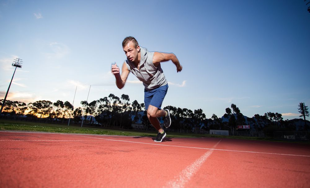 Man sprinting on a red track, blue shorts, gray top, under a clear sky, sunset in background.