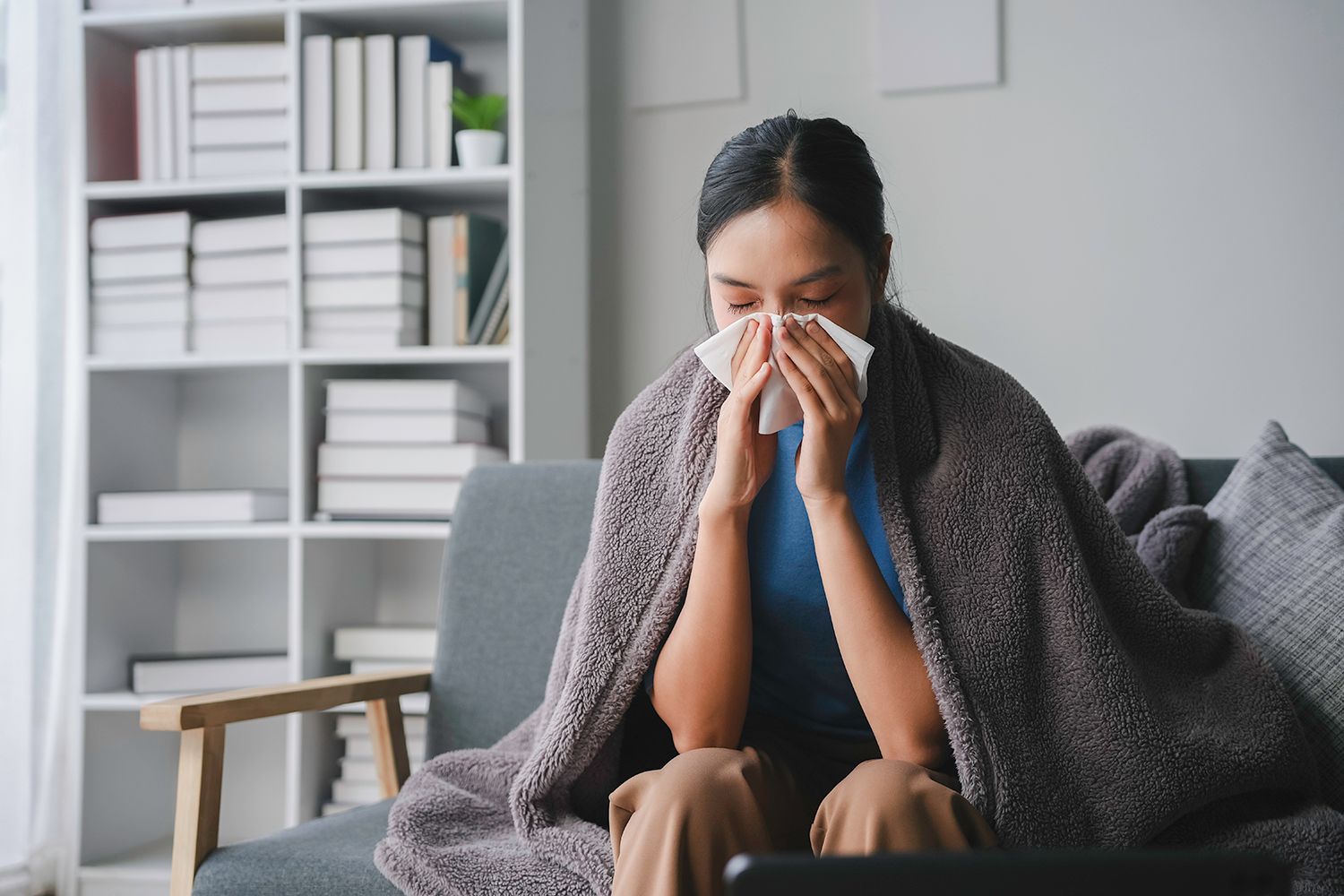 Person sitting on a couch, wrapped in a blanket and blowing their nose with a tissue.