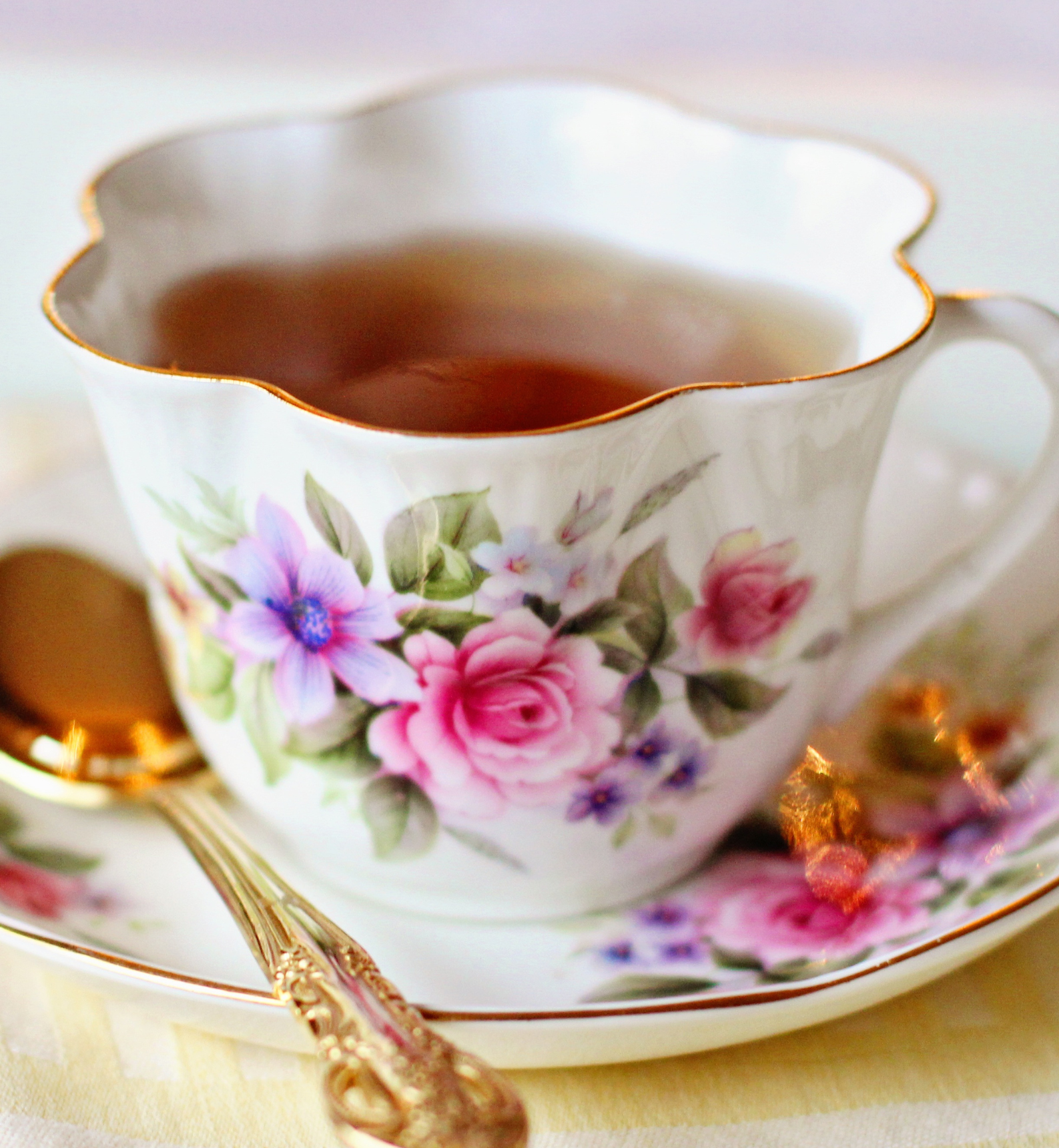 A cup of tea with flowers on it sits on a saucer next to a spoon