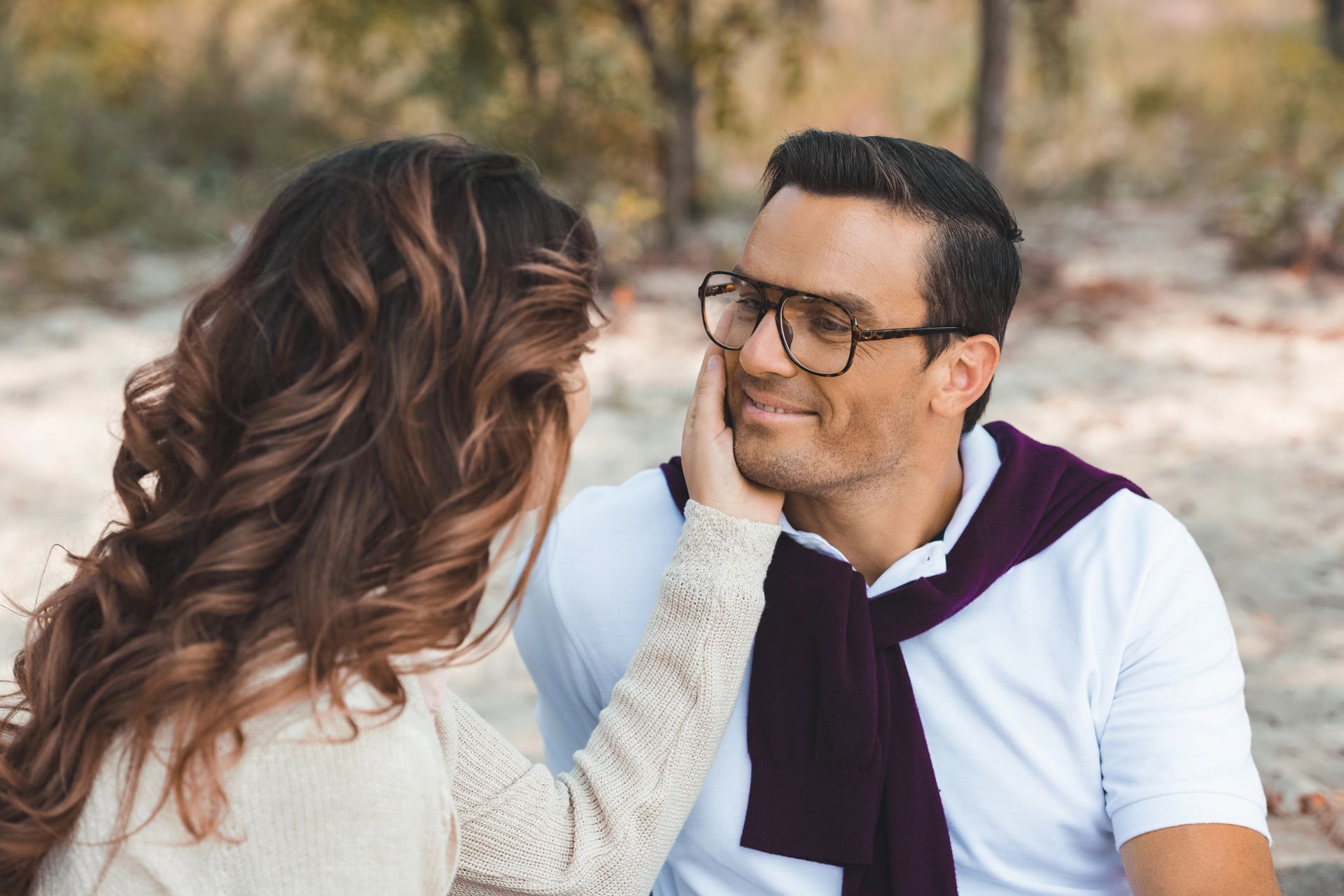 Una donna accarezza il volto sorridente di un uomo all'aperto; lui indossa occhiali e una camicia bianca con un maglione viola appoggiato sulle spalle.
