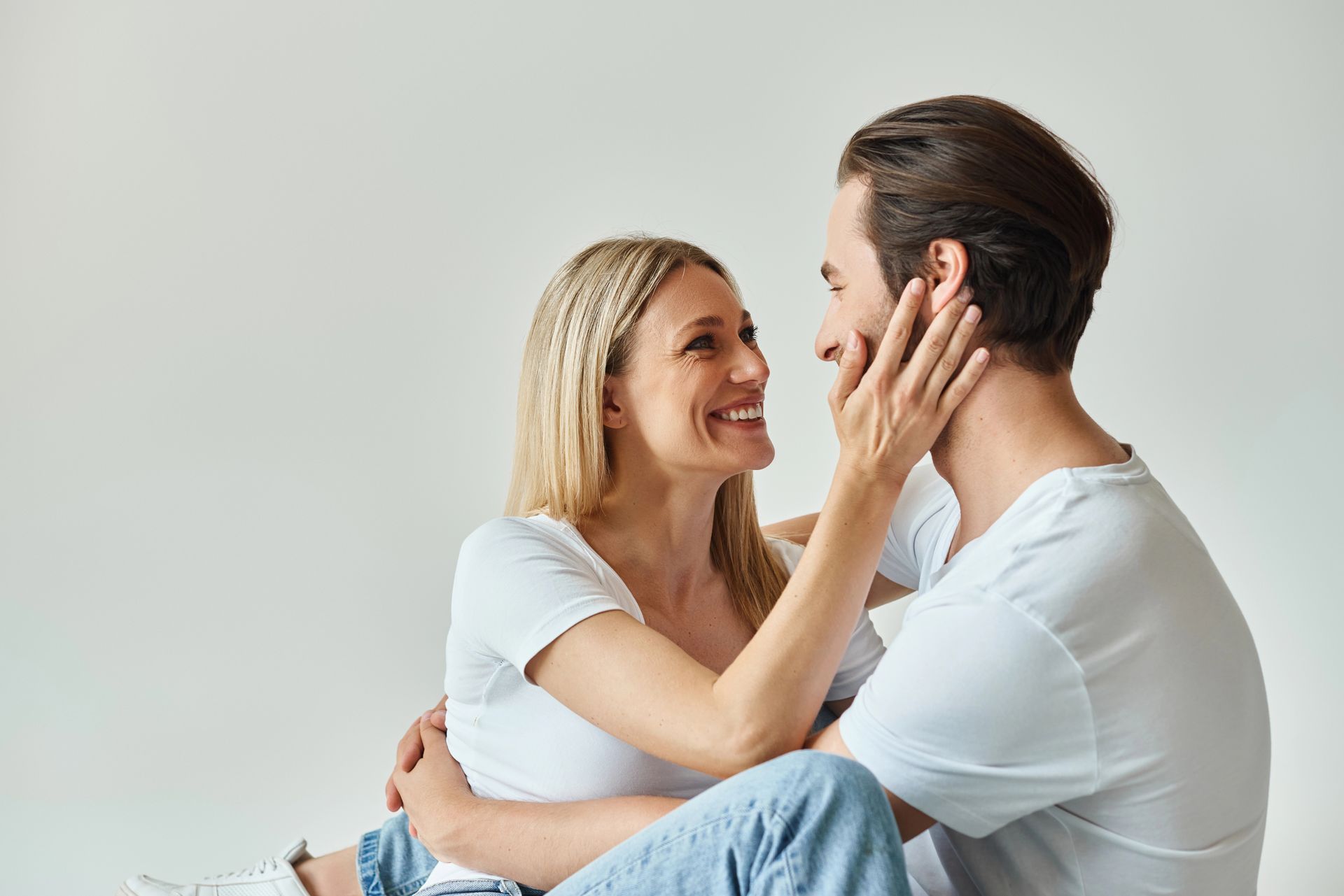 Una coppia seduta vicina su un letto, sorride e si accarezza il viso su uno sfondo neutro.
