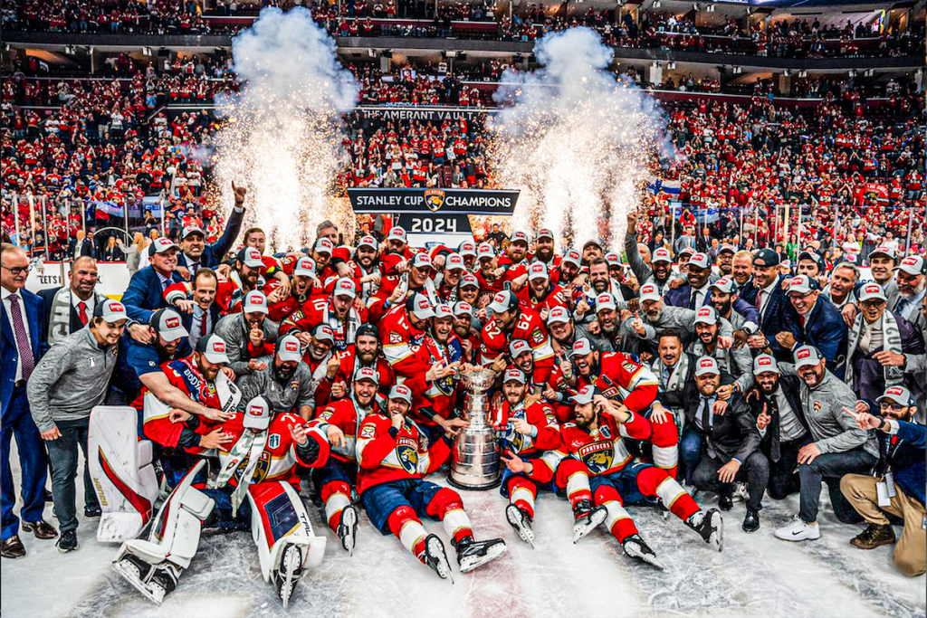 Hockey team in red and white uniforms celebrate with trophy, fireworks; in arena.