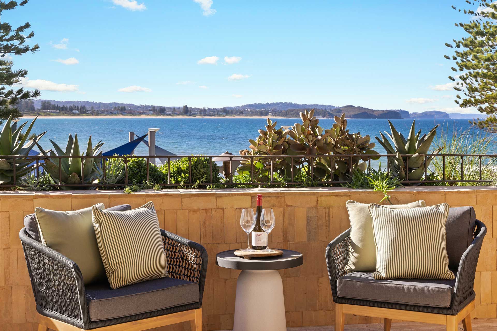 A balcony with two chairs and a table with a bottle of wine on it overlooking the ocean. Collaroy.