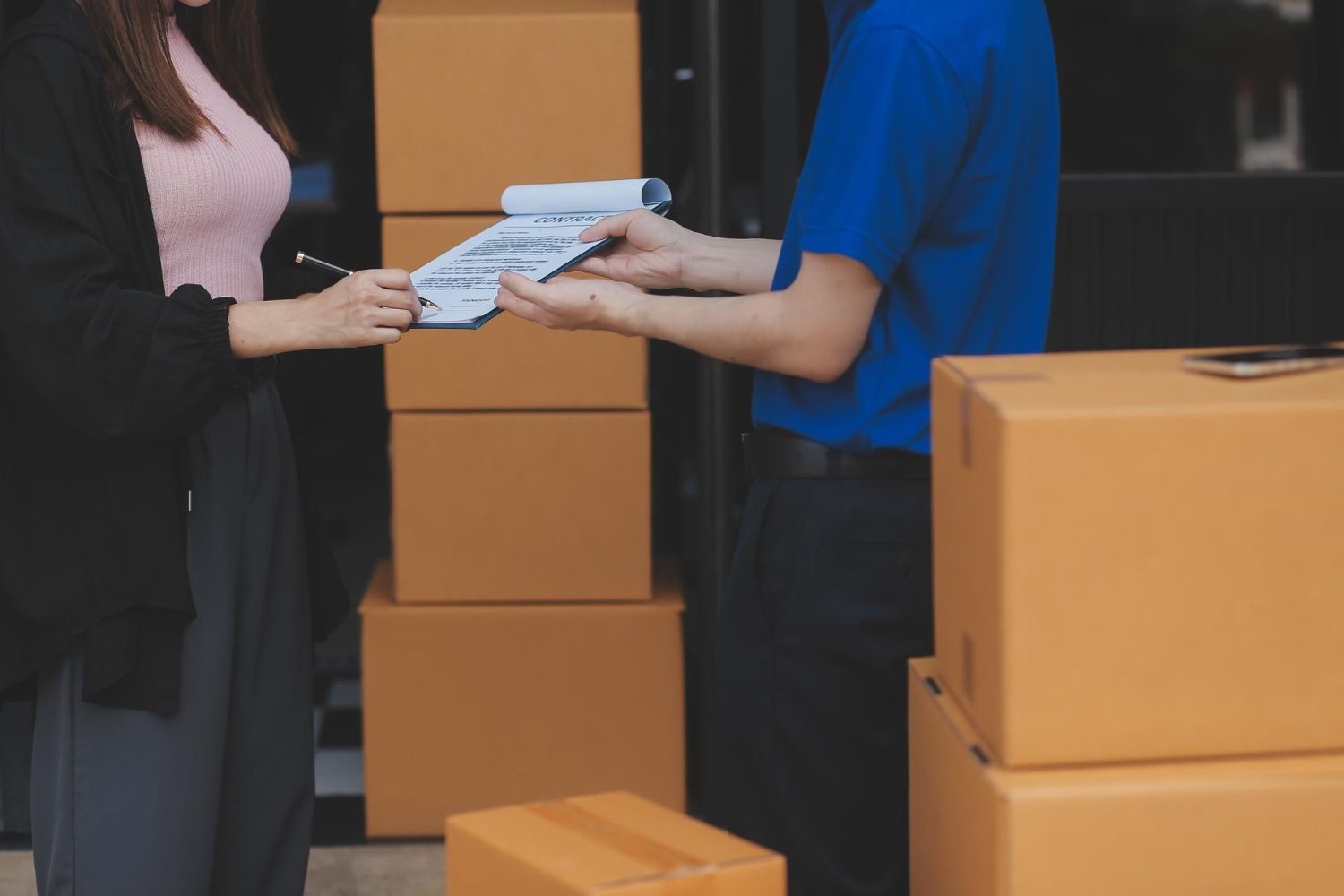 Two people exchanging papers beside stacked cardboard shipping boxes at a warehouse door