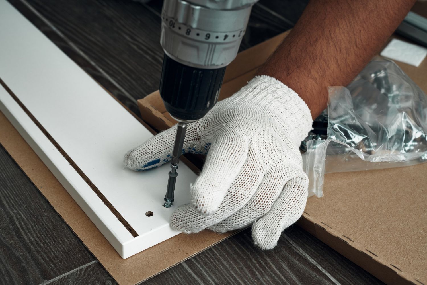 Gloved hand using a drill to assemble a white cabinet panel with screws on a tabletop
