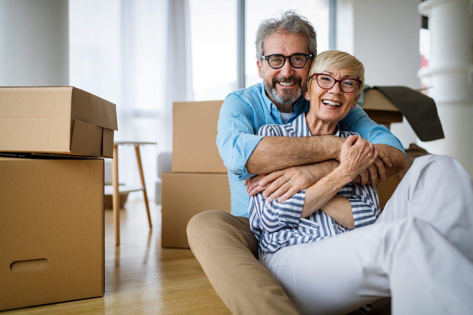 Couple smiling and hugging on the floor surrounded by moving boxes in a new home.