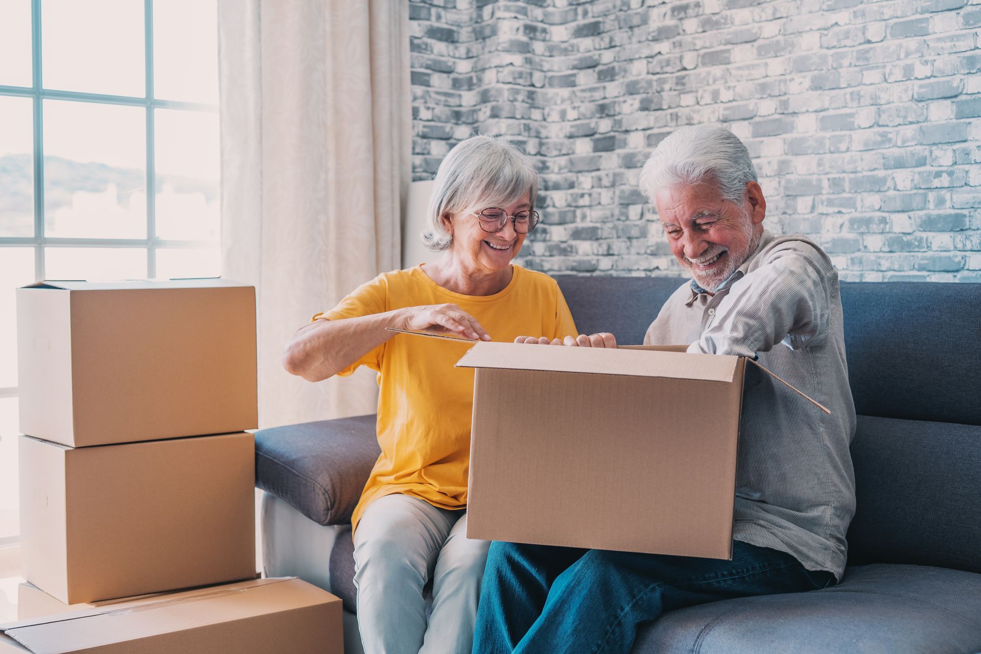 Two people sit on a couch in a bright room, smiling as they unpack cardboard moving boxes together.