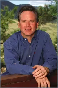 Man in a blue shirt leaning on a railing outdoors with a blurred mountain and trees in the background