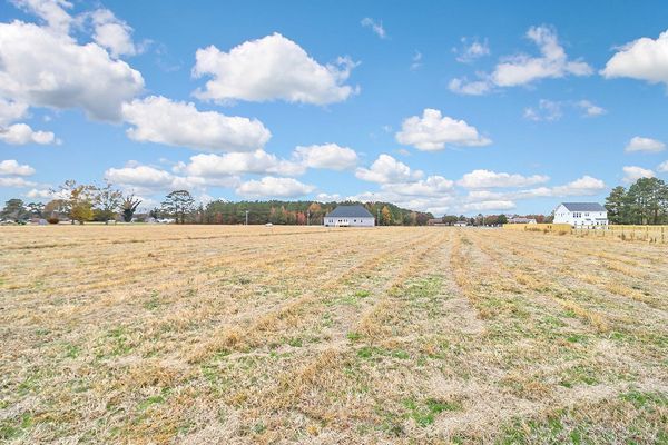 Open field with dry grass under a bright blue sky with scattered clouds, two houses visible in the distance.