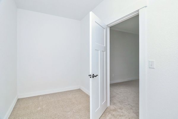 White door open to a small room with beige carpet and walls; another room visible in the background.