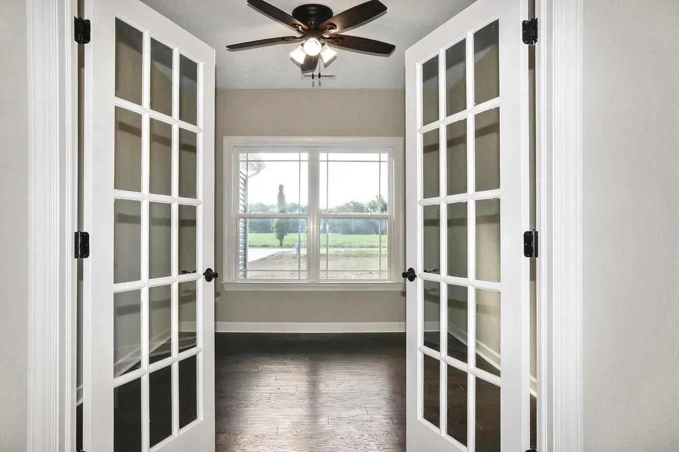 French doors open to a room with a window overlooking a field, dark wooden floor, neutral walls.