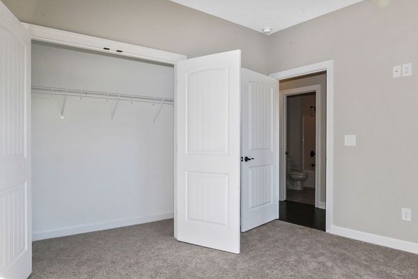 Empty bedroom with open closet, door, and doorway to a bathroom. Beige walls, gray carpet.
