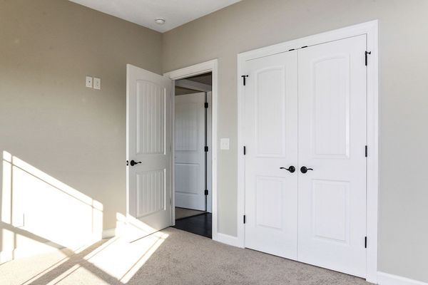 Empty room with beige walls, white doors, and gray carpet. Sunlight streams in.