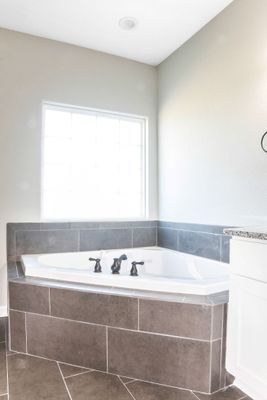 Corner bathtub with grey tile surround under a window in a neutral-toned bathroom.