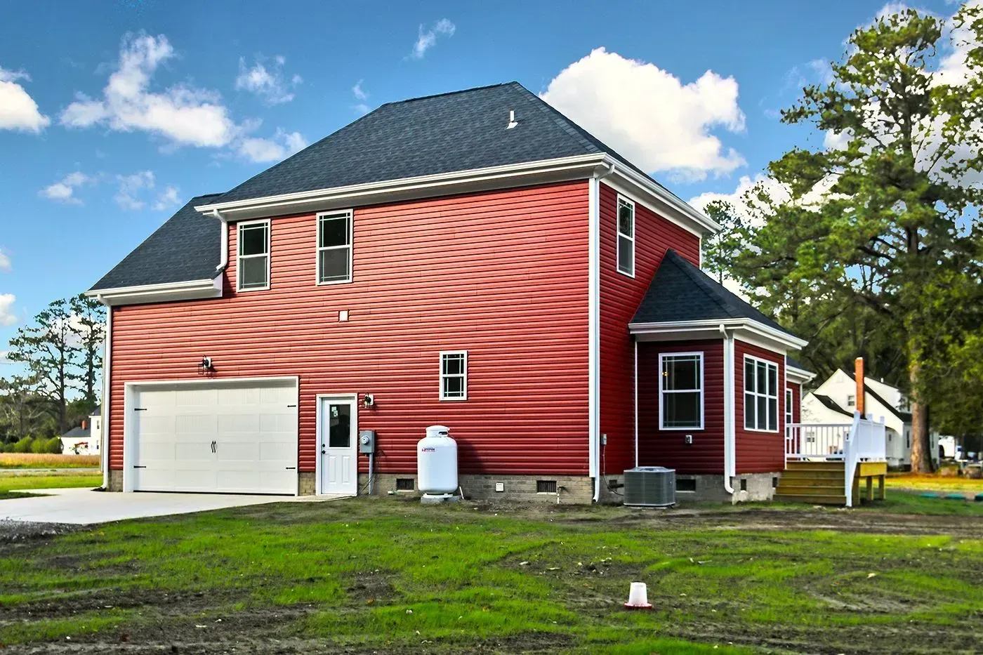 Red two-story house with a white garage door, set in a grassy yard under a blue sky.