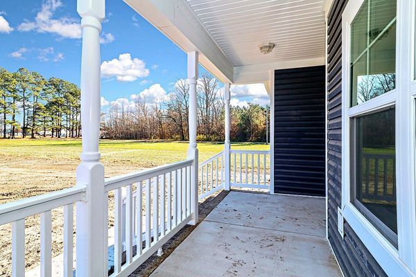 Covered porch with white columns and railings, concrete floor, overlooking a grassy field under a cloudy sky.
