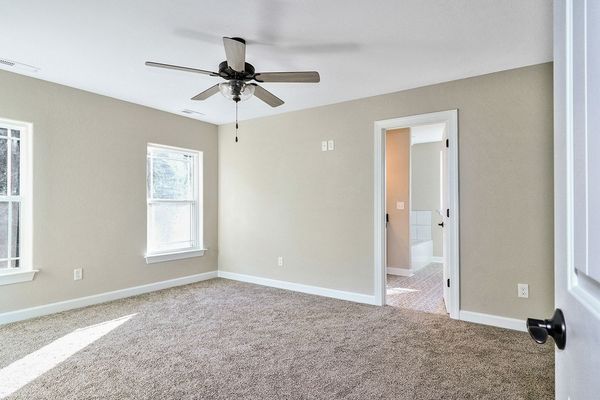 Empty bedroom with beige walls, carpet, and a ceiling fan, with a doorway to another room.