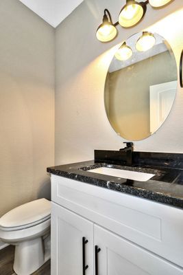 Bathroom with white vanity, black countertop, oval mirror, and toilet. Beige walls, light fixtures.